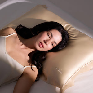 A woman with long dark hair sleeps peacefully on a bed, resting her head on the us.drowsysleepco Hyalo Pillowcase in Champagne Gold. She wears a cream-colored camisole and appears calm and relaxed.