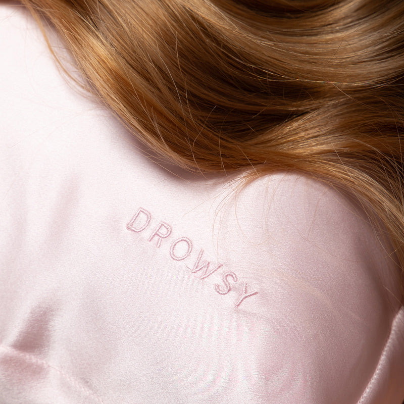 Close-up of long, light brown hair resting on a pale pink Hyalo Pillowcase - Petal Pink by us.drowsysleepco, with “DROWSY” embroidered in matching pink thread.