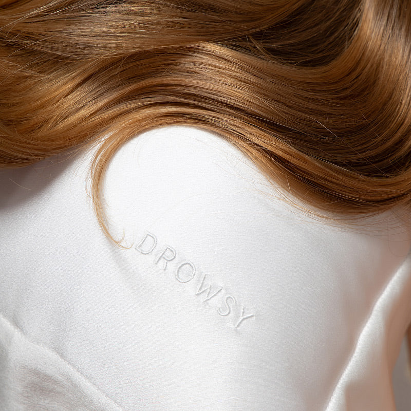 A close-up of long brown hair resting on the smooth, white Hyalo Pillowcase - Akoya Pearl by us.drowsysleepco, embroidered with the word DROWSY in subtle, raised letters.