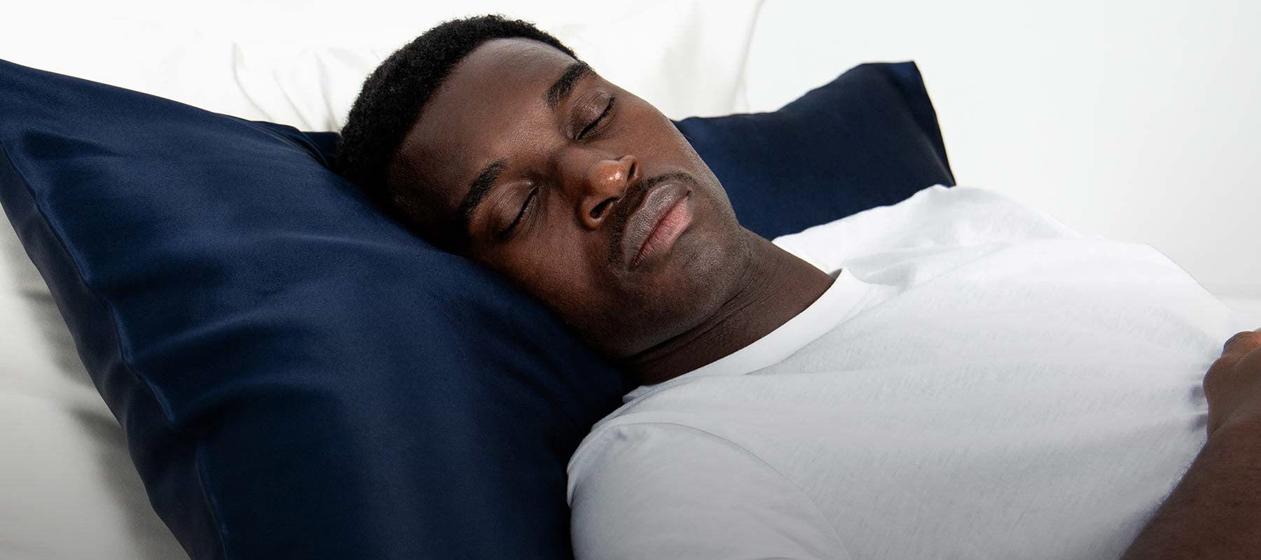 A man in a white shirt sleeps peacefully on a drowsysleepco Midnight ritual bundle pillowcase in Midnight Blue, surrounded by all-white bedding for a calm atmosphere.