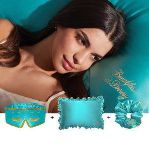 A woman with long dark hair rests on a turquoise satin pillow embroidered with "Breakfast at Drusy." In front is drowsysleepco’s Hollywood Experience Bundle: a turquoise silk sleep mask, ruffled pillow, and scrunchie for beauty sleep.