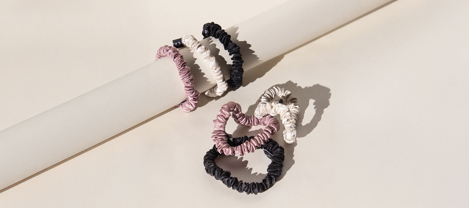 Three fabric scrunchies—one black, one cream, and one dusty pink—are looped over a white cylindrical rod, casting shadows on a beige background. Two more scrunchies lie on the surface below.