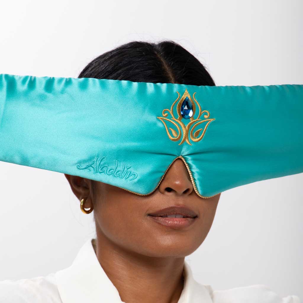 A woman wears the drowsysleepco Jasmine Jewel Sleep Mask—teal satin silk with gold embroidery and a blue jewel design—paired with gold hoop earrings and a white collared shirt, set against a plain background.