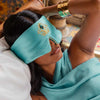 A woman relaxes on a bed wearing the Jasmine Jewel Sleep Mask by drowsysleepco, an Aladdin-inspired turquoise silk mask that matches her outfit. She rests one arm on her head, gold bangles adorning her wrist.