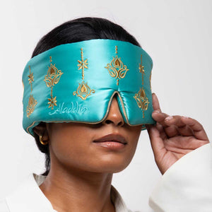 A person wears the drowsysleepco Jasmine Sleep Mask, a turquoise satin eye mask with gold embroidery, slightly lifting one side. The background is plain white.