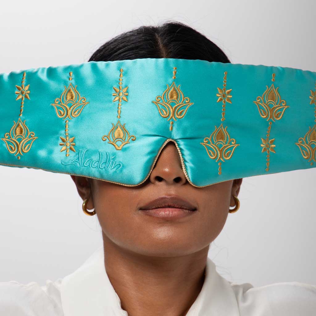 A woman in gold hoop earrings and a white shirt wears the drowsysleepco Jasmine Sleep Mask—turquoise with gold embroidery, ornate patterns, and 'Aladdin' stitched on it.