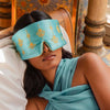A woman in a blue outfit relaxes on a pillow, wearing the drowsysleepco Jasmine Sleep Mask—an Aladdin-inspired blue and gold eye mask—in an ornate, colorful room with patterned walls and columns.