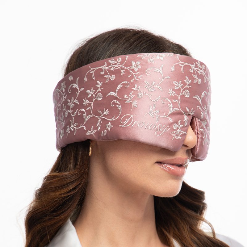 A person wears a dusty rose mulberry silk Pride & Prejudice - Sage + English Rose Sleep Mask embroidered with white floral vines and the word Drowsy.