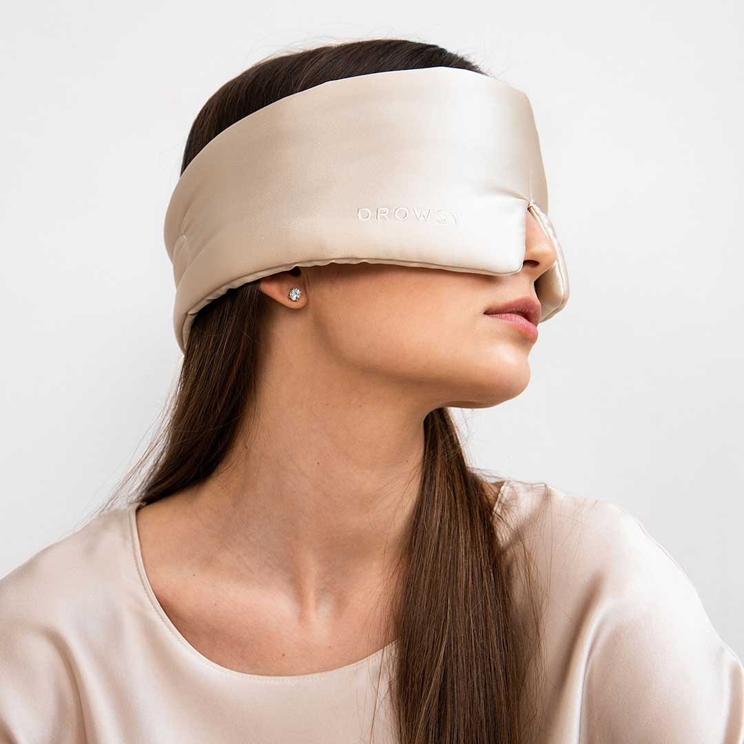 A woman with long brown hair wears the drowsysleepco Sensual sleep bundle in Dusty Gold—a large silk mask covering her eyes and ears, paired with a matching satin blouse—facing sideways against a plain white background.