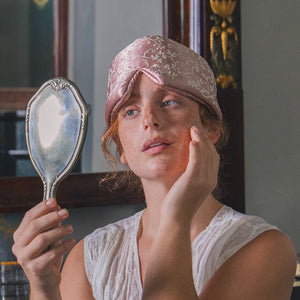 A person wearing a pale pink floral Pride & Prejudice - English Rose Sleep Mask on their forehead while looking into a handheld mirror.