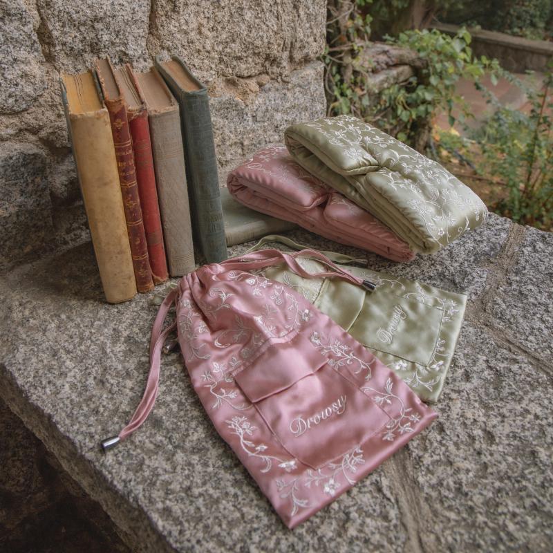 Two Pride & Prejudice - Complete collection original mulberry silk sleep masks and their matching drawstring pouches resting on a stone ledge next to a row of antique books.