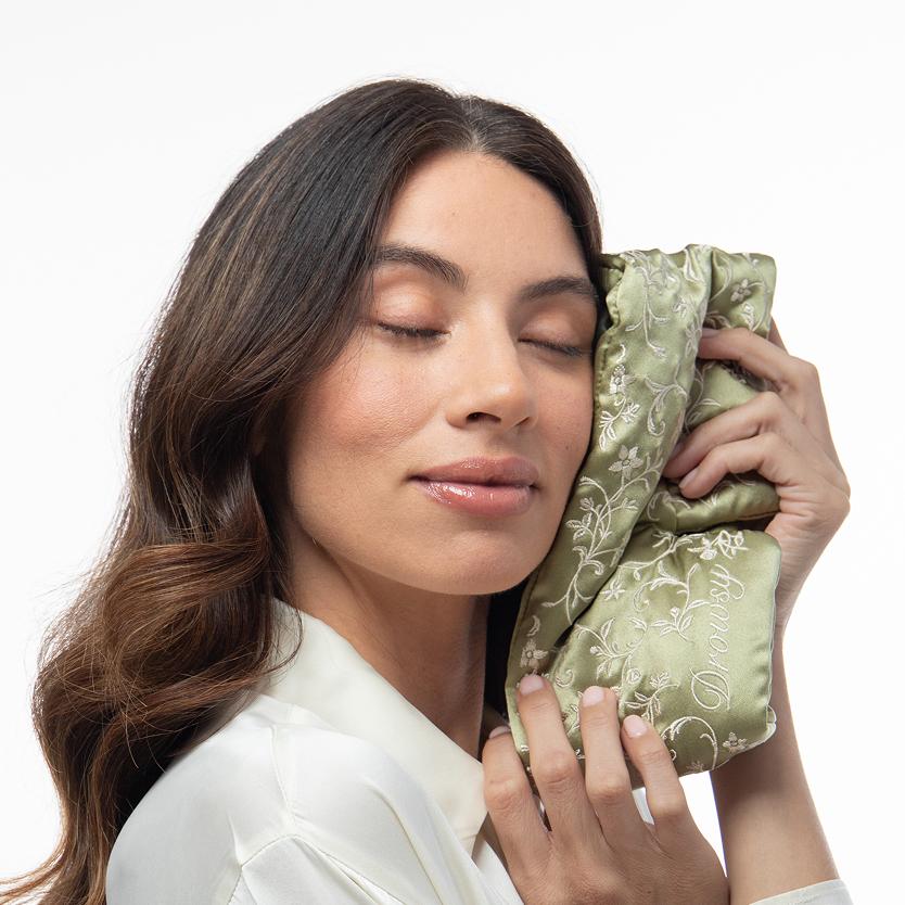 A woman with her eyes closed holds a sage green Pride & Prejudice - Sage + English Rose Sleep Mask with white floral embroidery against her cheek.