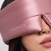 Close-up of a woman wearing the Damask Rose silk sleep mask by drowsysleepco, featuring a Swarovski®-embellished logo on the side. The soft pink mask comfortably covers her eyes and part of her nose, ensuring undisturbed beauty rest.