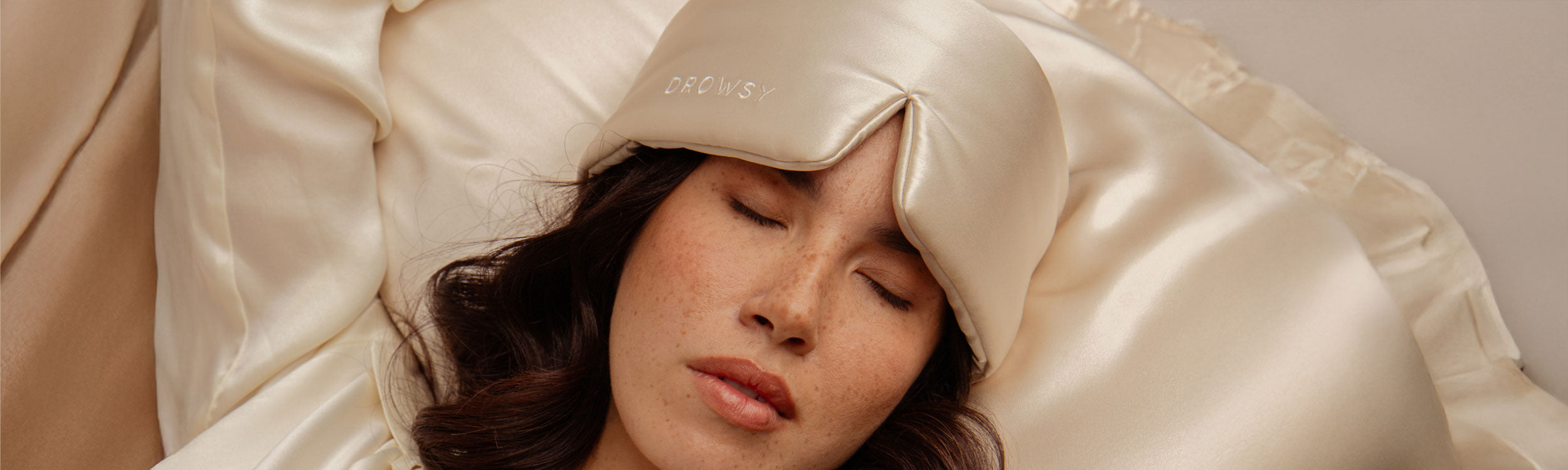 A woman with closed eyes lies on cream-colored satin bedding, wearing a matching satin sleep mask with DROWSY written on it. Her dark hair is visible, and the scene suggests peaceful rest or sleep.