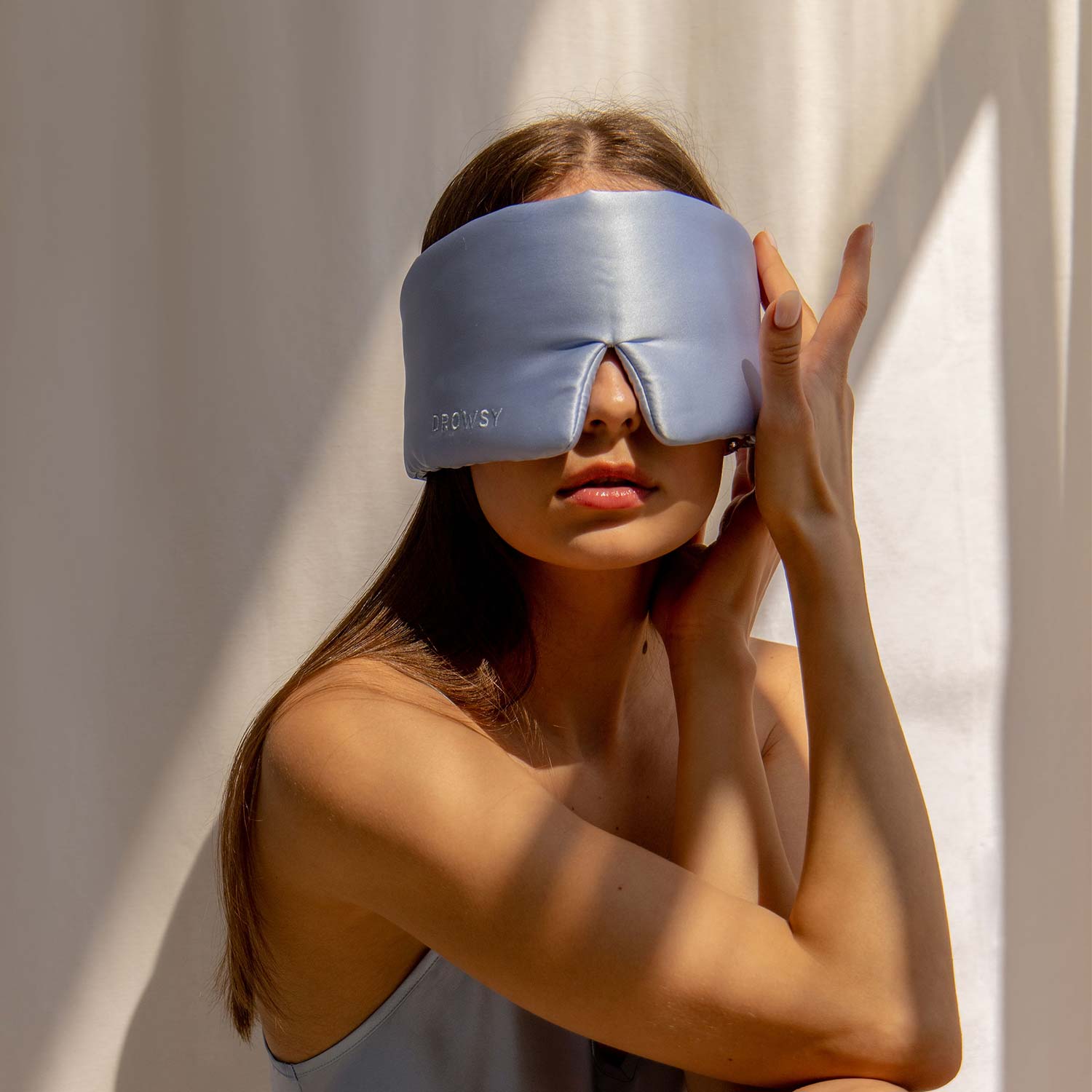 Model wearing baby blue coloured silk sleep mask in the sunlight with a white background