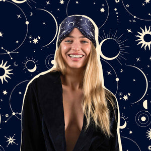 A smiling woman with long blonde hair wears drowsysleepcos Cosmic Energy celestial headscarf and black robe against a dark blue cosmos-themed backdrop—ideal inspiration for restful nights or picking the perfect silk sleep mask.